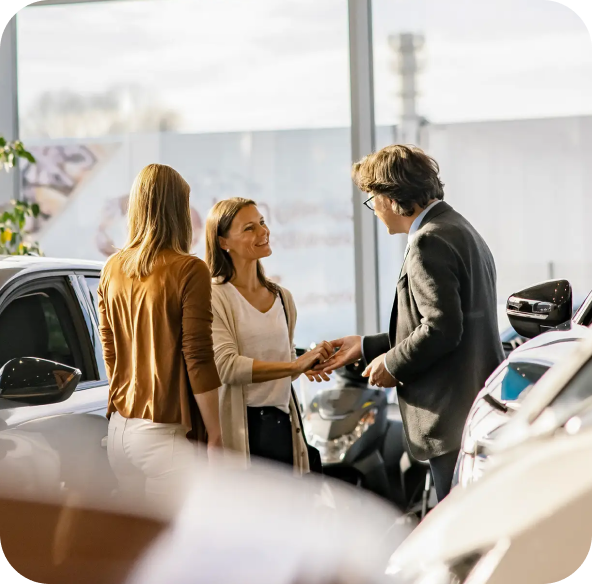 Two women speaking with a salesperson inside a sunlit car dealership, surrounded by modern vehicles and large windows