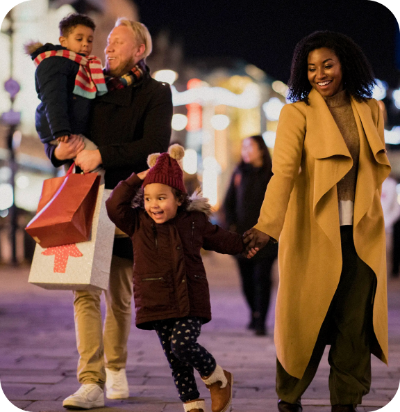 A family strolls through a festive shopping district at night, bags in hand and smiles on their faces—capturing the joy of thoughtful purchases and time well spent together.