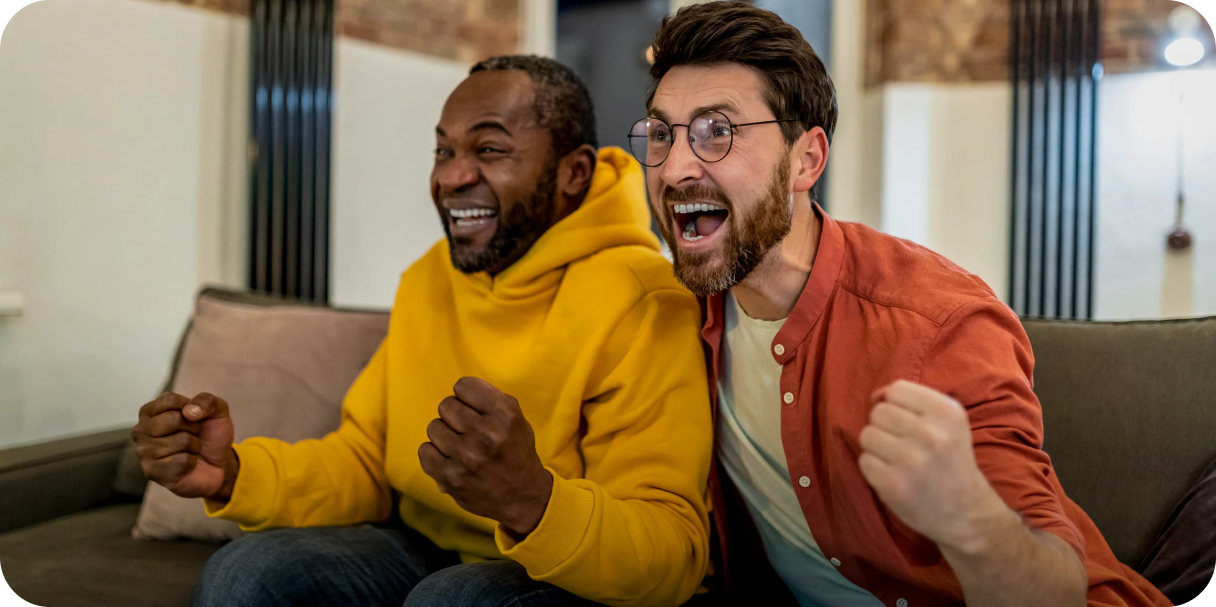Two men sitting on a couch with enthusiastic expressions, celebrating while looking at a screen, suggesting a shared moment of success or excitement.