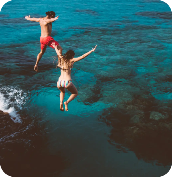 Two people leap off a rocky cliff into clear turquoise water, capturing a carefree and adventurous travel experience. 