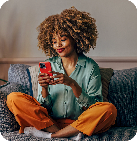 A woman sitting comfortably on a couch, smiling while using a smartphone—capturing a relaxed moment of enjoying perks or rewards with ease.