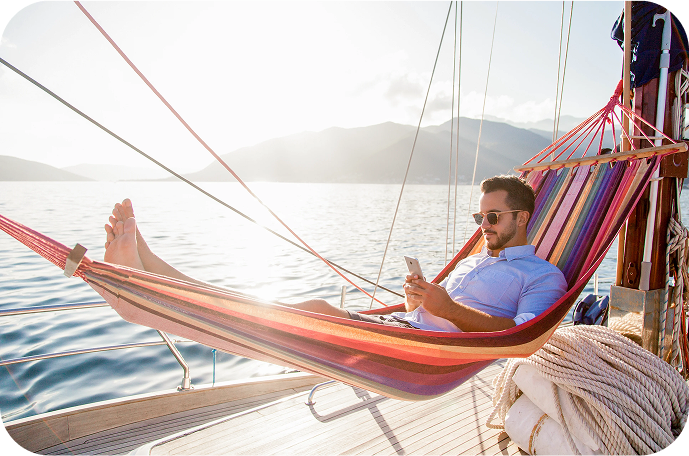 Man relaxing in a hammock on a sailboat, looking at his phone with mountains and water in the background.