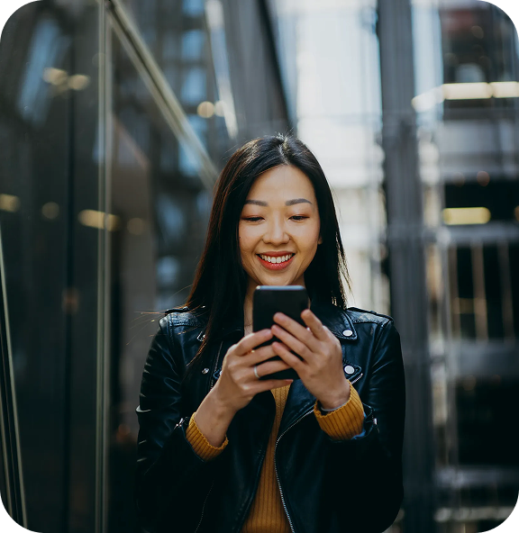 A woman in a street setting looking down at her smart phone and smiling