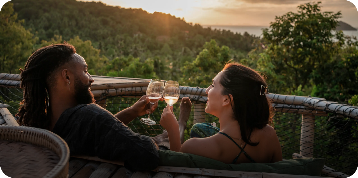 A couple relaxing in a hammock-style lounge and toasting glasses at sunset, overlooking a lush tropical landscape—capturing the ease and joy of travel