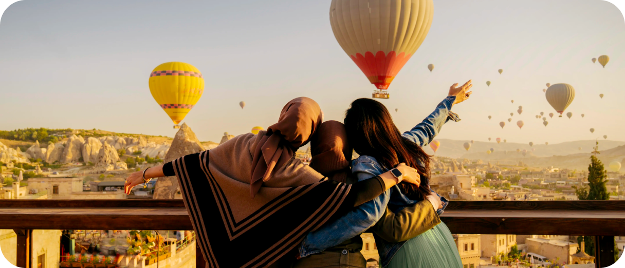 Three women with their arms around one another face the opposite direction while watching dozens of hot air balloons take off in the distance